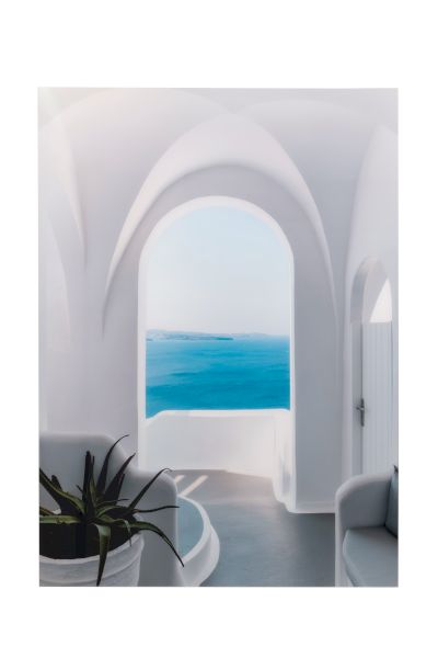 A bright, airy room with smooth white arches opens to a blue sea beyond a curved doorway, a potted plant on the left and cozy seating nearby.