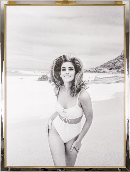A woman in a white bikini posing on a beach with rocks in the background, framed by a gold border.