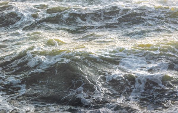 Turbulent ocean waves churn with foamy white crests and dark green-blue depths, catching light in rippling, swirling motion.