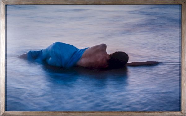 A man lies face down on calm water, head turned, wearing blue shorts; a serene, blue-toned scene in a wooden frame.