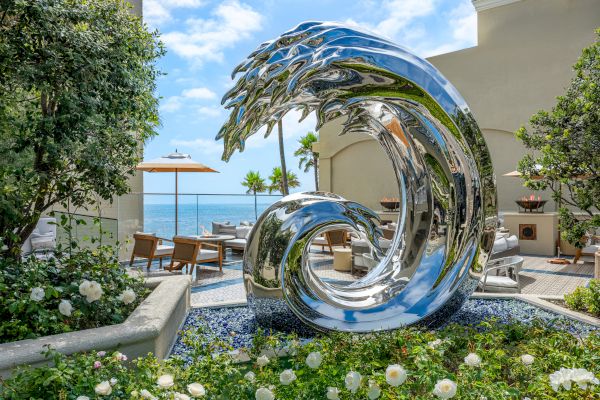 A shiny abstract sculpture curls in a circular, reflective loop by a pool, with lounge chairs, greenery, and a sunny ocean view in the background.