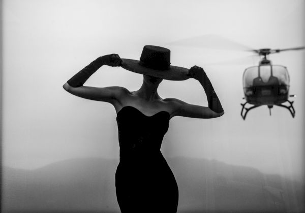 A stylish woman in a strapless dress adjusts a wide-brim hat, with a helicopter in the background against a hazy landscape. (140 chars)