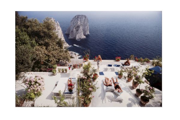 A sunny terrace with white stone loungers and potted plants overlooks a blue sea and rocky island cliffs in the distance.