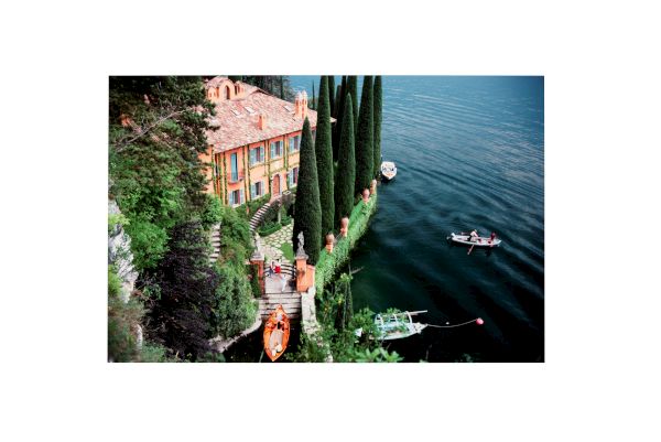 A scenic lakeside villa with tall cypress trees lines the water, boats docked by a stone terrace, and a calm blue lake with a few boats.